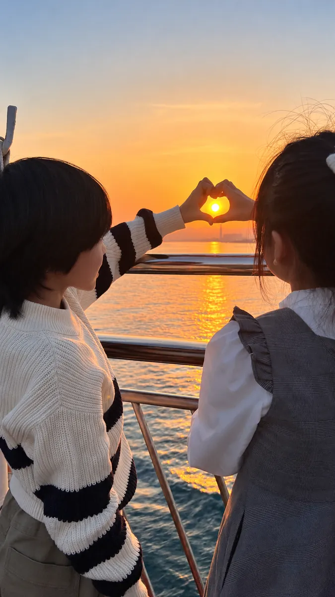 船の上から、夕日を手のハートで囲む子どもたち